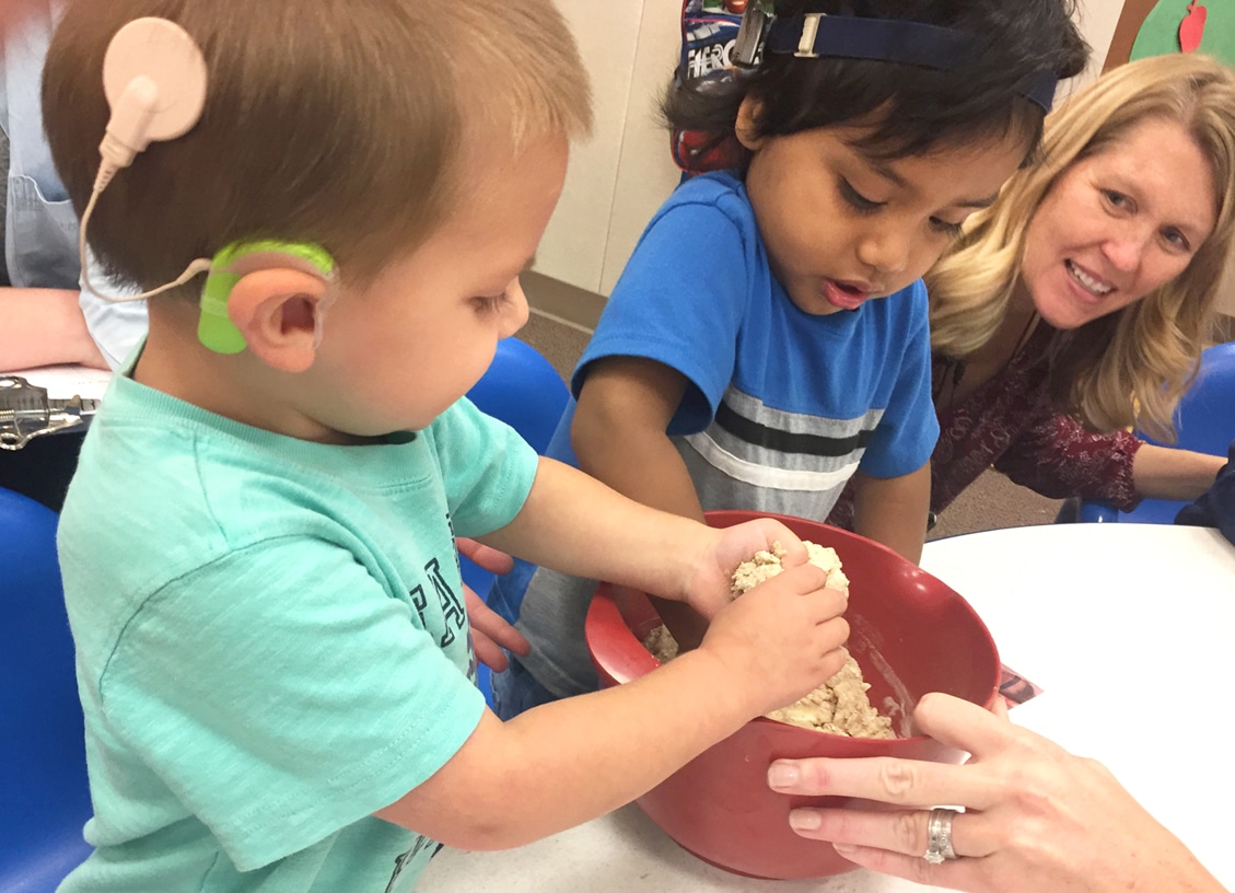 two toddlers, one with a cochlear implant, stir dough in a bowl while teacher looks on