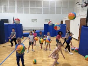 A bunch of kids are throwing beachballs in a gym.